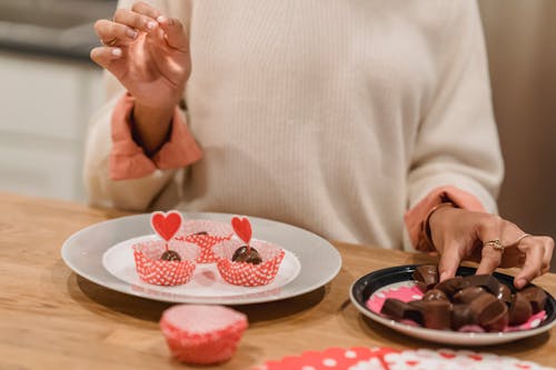 Unrecognizable female serving sweet chocolate candies into molds on plate decorated with red hearts while standing near table in kitchen at home