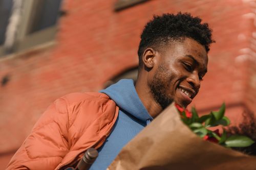 Free From below of young African American male in stylish outfit carrying flowers wrapped in craft paper on street looking away Stock Photo