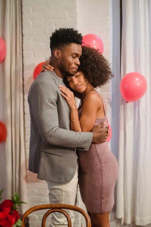 Free Side view of dreamy young black couple in elegant clothes standing in bright room and cuddling while dancing together with closed eyes near pink balloons on wall Stock Photo