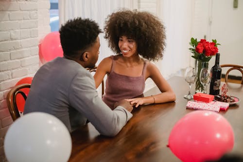 Free Young black couple in elegant clothes talking while sitting at table with wine bottle and glasses near flowers bouquet in vase and balloons in home while looking at each other Stock Photo