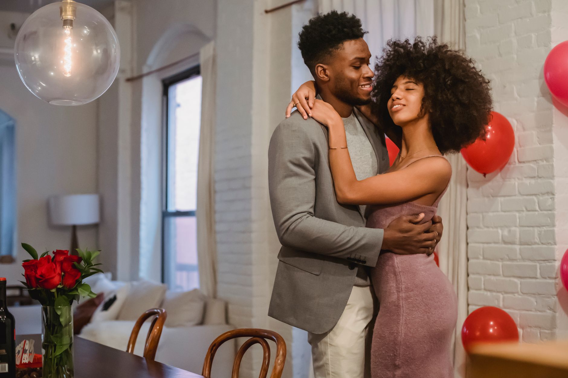 Smiling young African American couple in elegant outfits embracing and dancing while standing in bright apartment near balloons on wall and table with bouquet of roses in vase