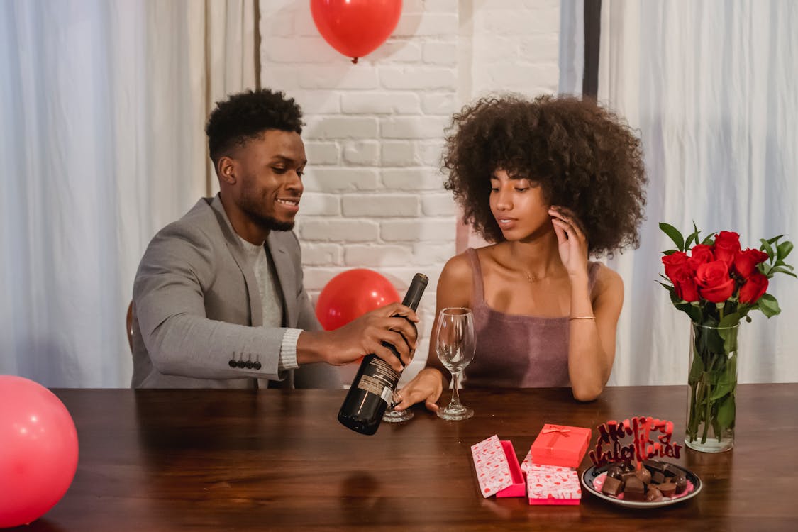 Free Young black couple in elegant clothes sitting at table while male pouring red wine from bottle in glasses near gifts and vase with flower bouquet and chocolates in light loft Stock Photo