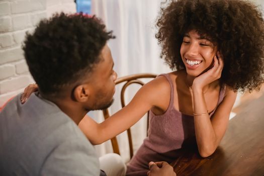 Positive young African American couple in elegant outfits in light room sitting at table in chairs and having conversation while looking at each other near curtains