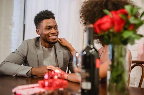 Free Happy young black couple in elegant clothes talking while holding hands at table with wine bottle and glasses near flowers bouquet in vase at home while looking at each other Stock Photo