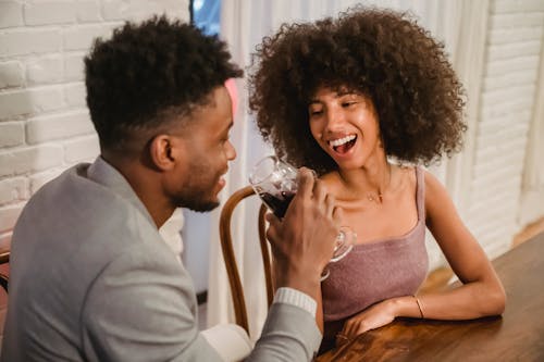Free Happy young African American couple in elegant clothes drinking red wine from glasses while sitting at table in bright apartment near curtains and brick wall Stock Photo