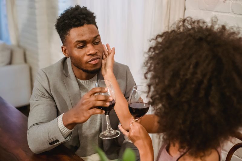 Young African American couple drinking red wine and looking at each other while sitting at table during romantic dinner in apartment