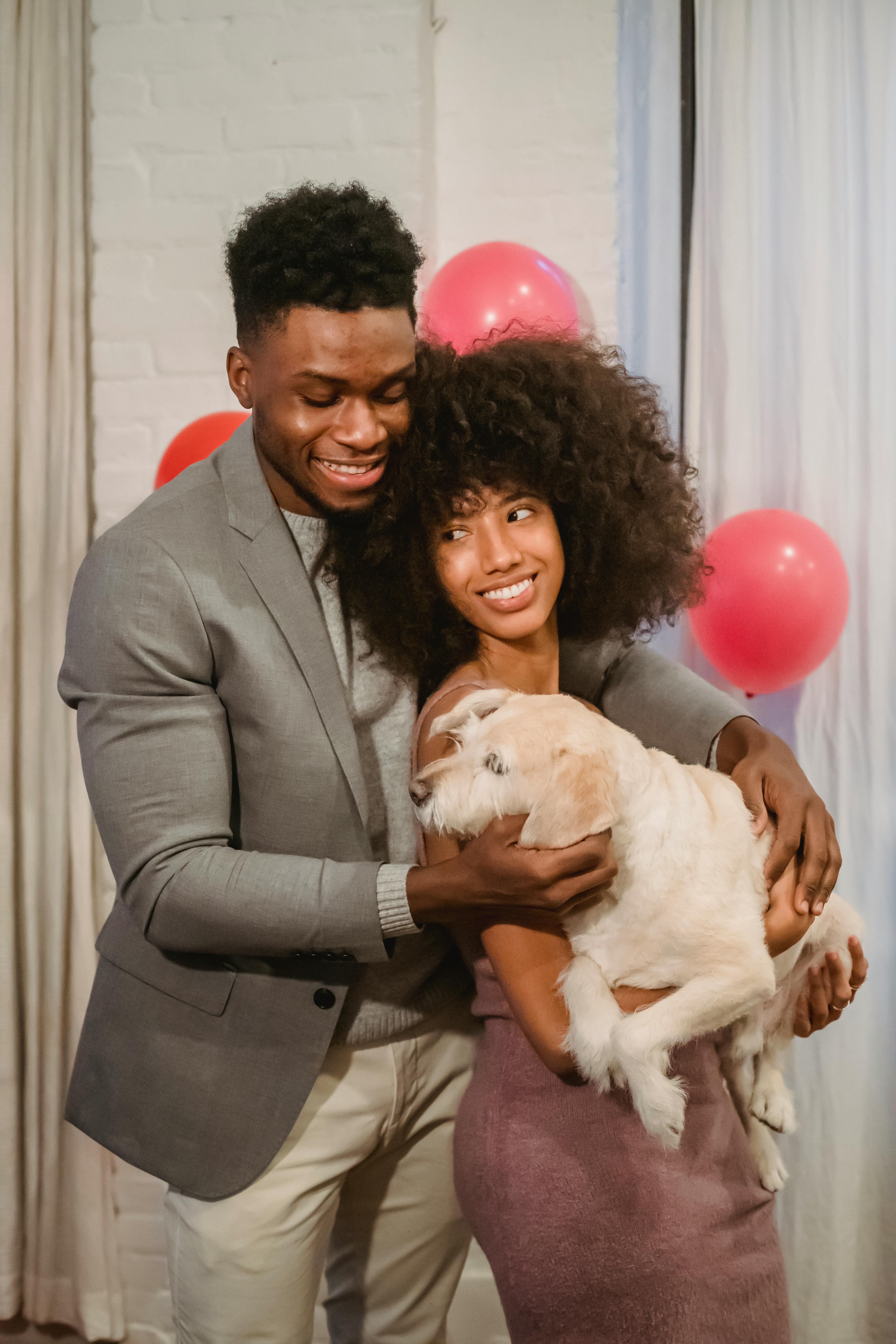 Cheerful African American couple in casual clothes embracing and stroking adorable dog while standing in decorated room
