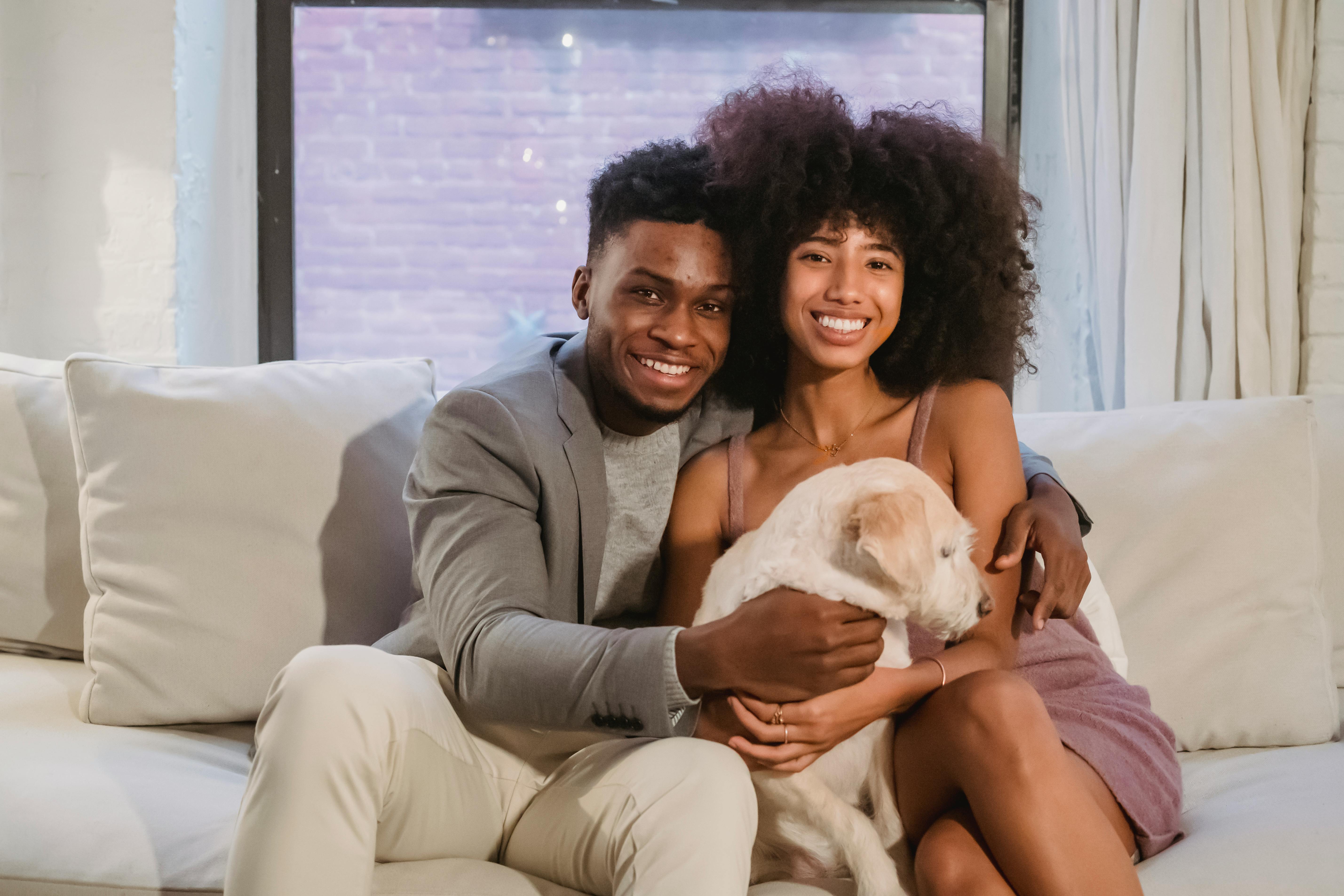 Happy young Black couple sitting on comfortable sofa with cushions and stroking adorable dog while spending time together in living room