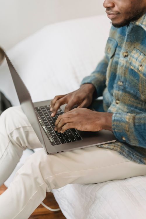 Free From above of crop concentrated African American male freelancer in casual clothes working remotely on laptop while sitting on comfortable bed at home Stock Photo