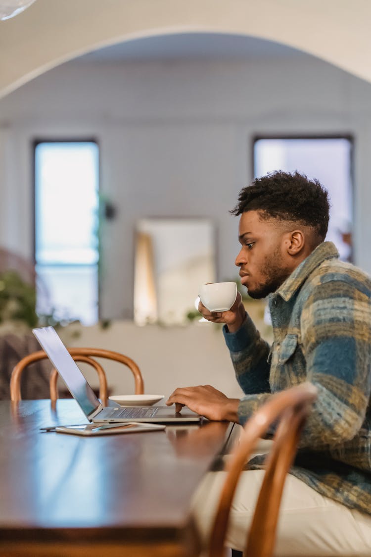 Serious Young Black Man Drinking Coffee While Working Remotely On Netbook At Home