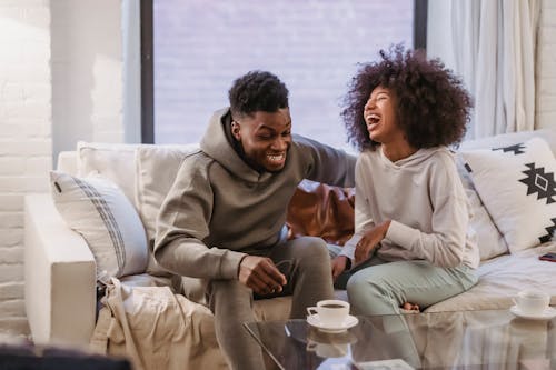 Free Cheerful young African American couple in casual outfit laughing happily while resting together on comfortable sofa at home Stock Photo