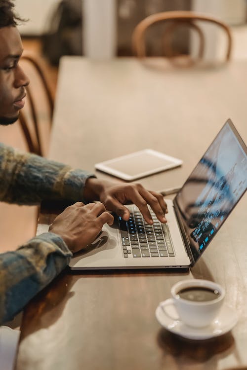 Free Side view crop of focused young self employed African American man typing on laptop while working remotely sitting at table with coffee cup at home Stock Photo
