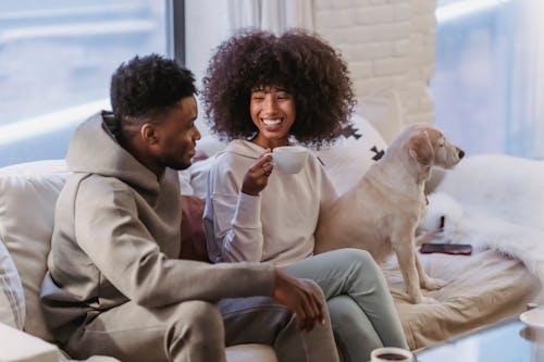 Free Cheerful young African American couple in stylish hoodies communicating and drinking coffee while resting on comfortable couch with cute bed Stock Photo