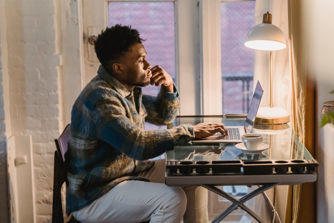 Side view of serious young bearded African American male freelancer in casual clothes typing on laptop sitting at table with cup of coffee during remote work at home