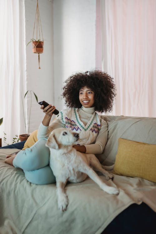 Happy young African American woman with curly hair switching channels on TV and smiling while resting on sofa with obedient dog