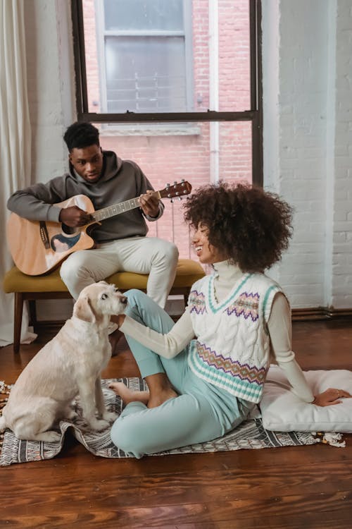Free Happy young ethnic woman stroking pet on floor near boyfriend playing guitar Stock Photo
