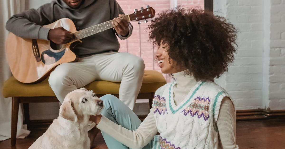 Happy young ethnic woman stroking pet on floor near boyfriend playing guitar