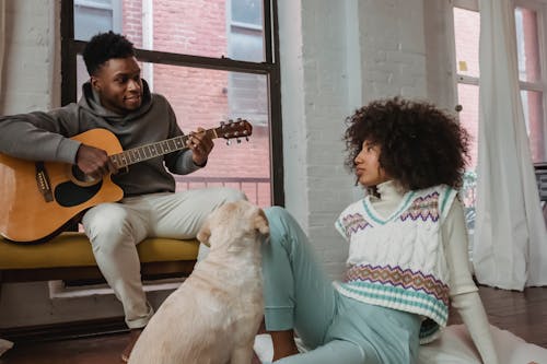 Cheerful black man playing guitar for girlfriend and dog at home