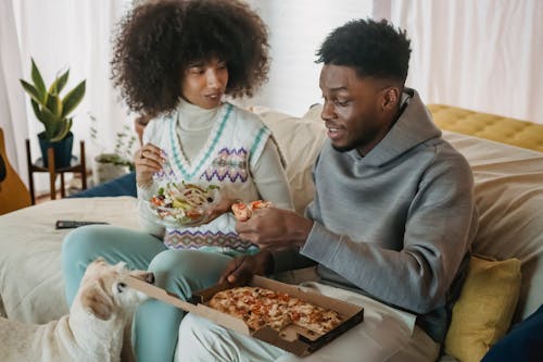 Positive African American couple eating salad and pizza while sitting on couch near cute dog in cozy living room at home