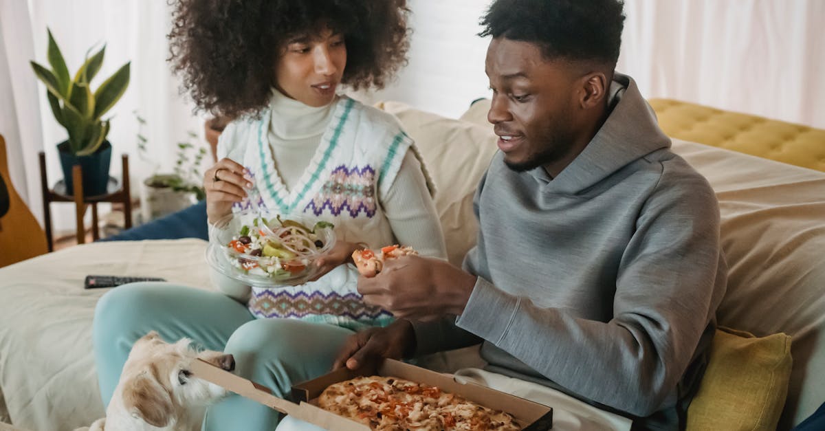 Positive African American couple eating salad and pizza while sitting on couch near cute dog in cozy living room at home