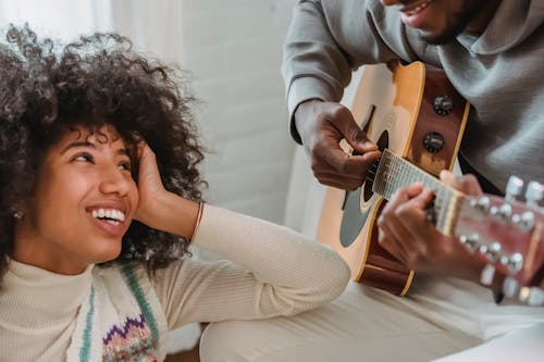 Unrecognizable African American male musician playing acoustic guitar for happy black girlfriend while spending time together in light room at home