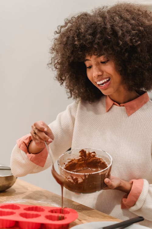 Positive African American female pouring chocolate batter from glass bowl into silicone molds while cooking pastry at table in kitchen