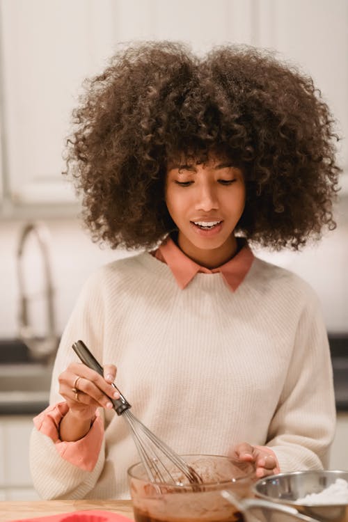 Positive African American female with whisk mixing ingredients for batter in bowl while cooking at table in kitchen on blurred background