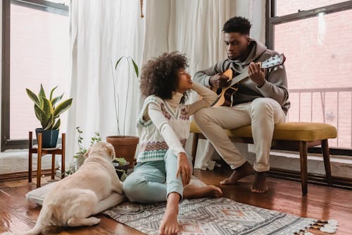 Free Full body of black boyfriend playing acoustic guitar for African American girlfriend while spending time together in room with dog Stock Photo