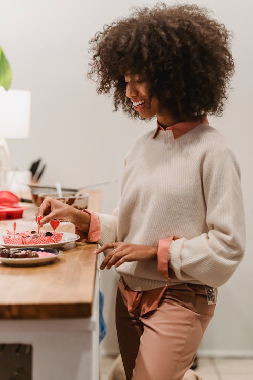Free Side view of happy African American female serving chocolate candies in molds with small hearts while standing at table in kitchen Stock Photo