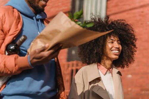 Free Cheerful African American woman strolling on sunny street near crop black boyfriend with bouquet of flowers and bottle of wine Stock Photo