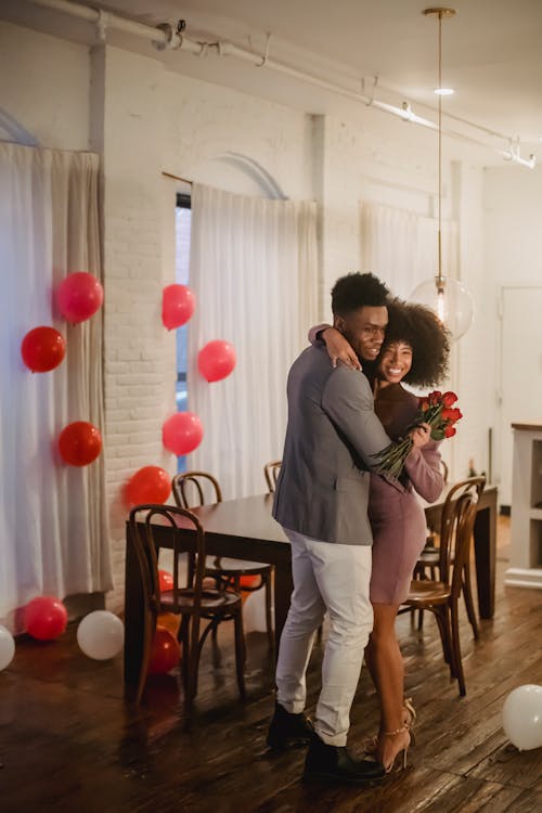 Full body of African American couple with bouquet of flowers embracing while standing in room with balloons during holiday celebration