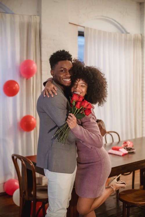 Free Cheerful African American couple with roses hugging and looking at camera while standing in room decorated with balloons during holiday celebration Stock Photo