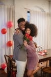 Free Cheerful African American couple with roses hugging and looking at camera while standing in room decorated with balloons during holiday celebration Stock Photo
