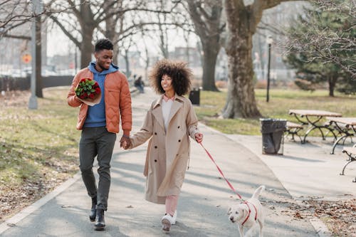 Full body of content African American couple with flowers holding hands while strolling on walkway with dog on leash during date