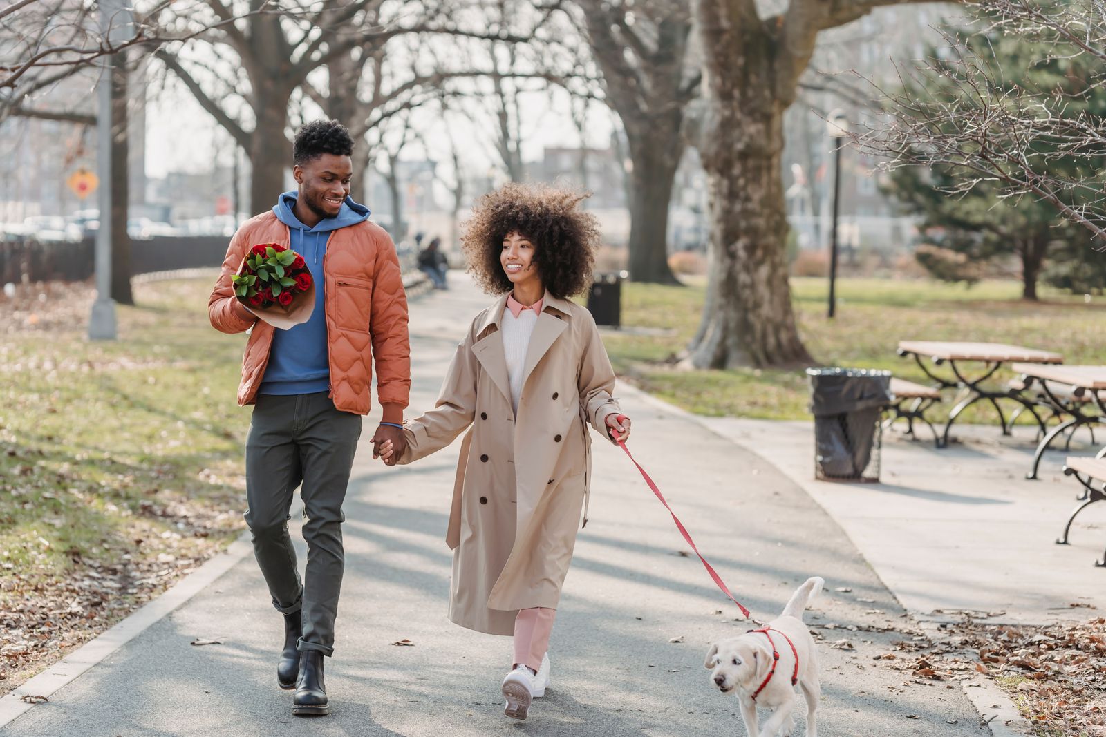 A smiling young Black couple holding hands as they walk a fluffy cream-coloured puppy on a red leash along a leaf-strewn park path on a sunny late-fall day