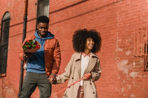 Free Cheerful African American girlfriend with leash and black boyfriend with bouquet of flowers holding hands while strolling on street Stock Photo