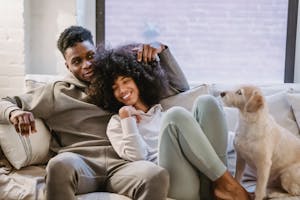 Cheerful African American couple in casual clothes cuddling on sofa near adorable dog while spending time together in living room