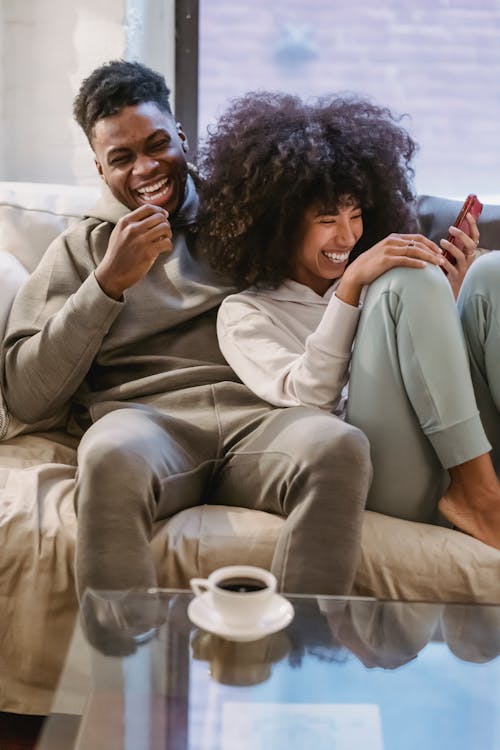 Positive African American couple in casual clothes browsing cellphone on sofa while having fun in cozy living room at home