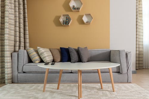 Modern design of living room in pastel colors furnished with big gray couch with cushions placed behind wooden oval table with white surface surrounded by dark yellow walls