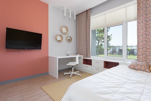 Modern design of comfortable bedroom with light workplace near big window and wide bed in opposite of TV set on coral wall