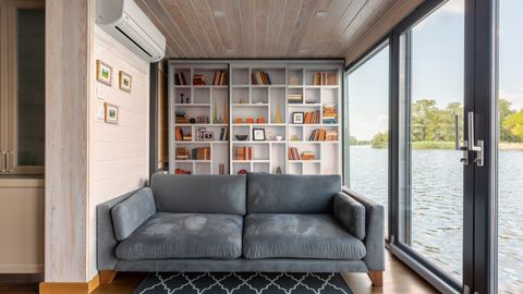 Interior design of modern house with majestic view on river surface through glass wall from comfortable soft gray couch placed in front of wide bookshelf