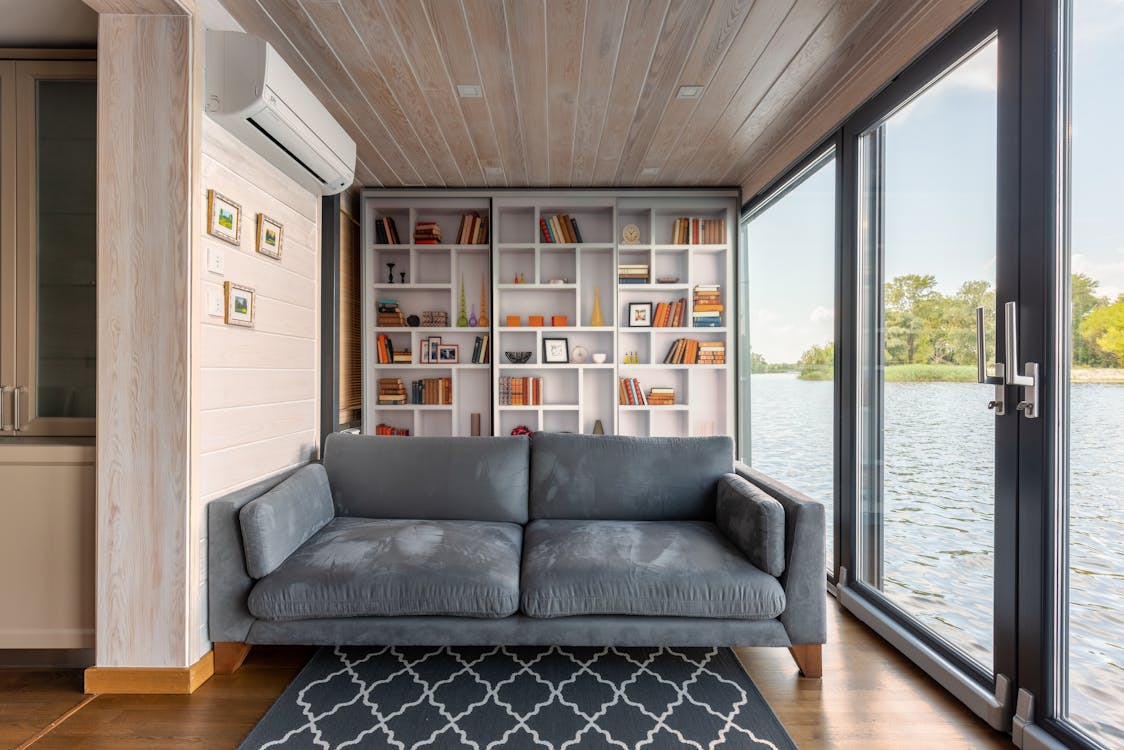 Free Interior design of modern house with majestic view on river surface through glass wall from comfortable soft gray couch placed in front of wide bookshelf Stock Photo