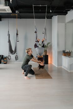 A yoga instructor guides a woman practicing aerial yoga inverted pose in a modern studio.