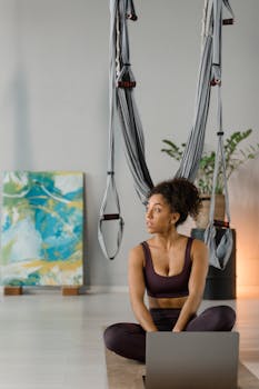 Woman practicing aerial yoga indoors with exercise hammock and laptop. Ideal for yoga and fitness concepts.