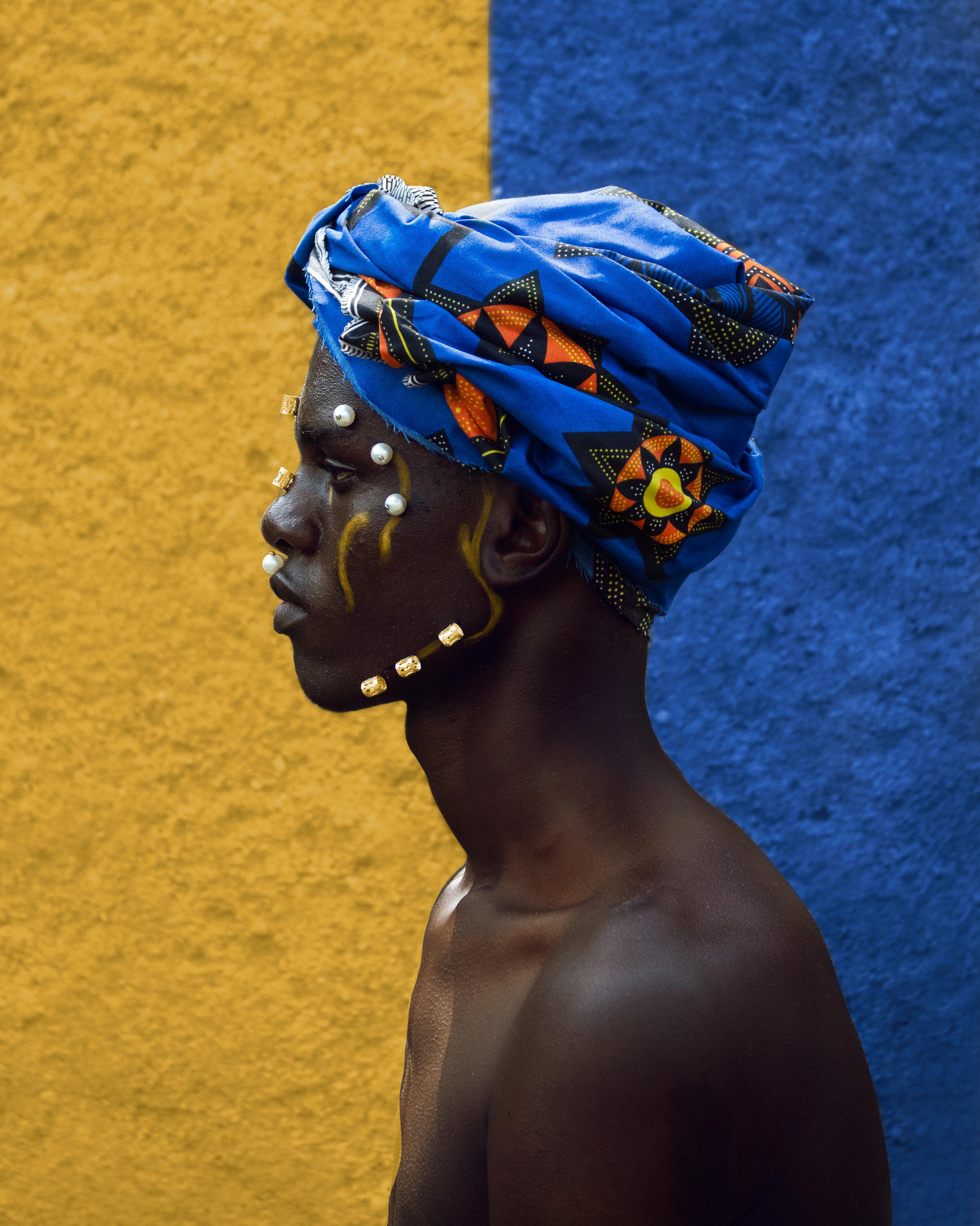 Artistic side view portrait of shirtless man in traditional attire with turban standing against a vibrant blue and yellow wall.