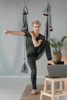 Adult man practicing aerial yoga at home, using a laptop for guidance. Focus on fitness and flexibility.