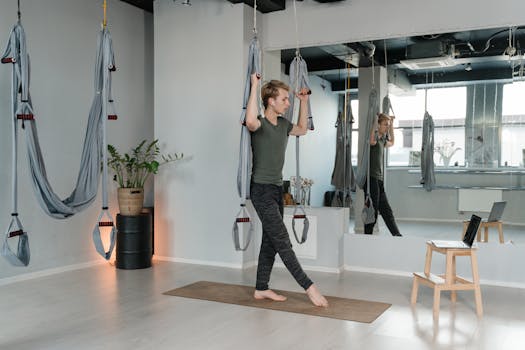 Adult man performing aerial yoga exercises indoors with hammocks and mirrors, focusing on flexibility and balance.