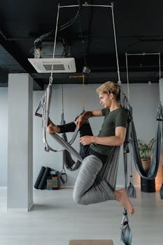 Adult man performing aerial yoga pose in an indoor studio, balancing on a hammock swing for flexibility and wellness.