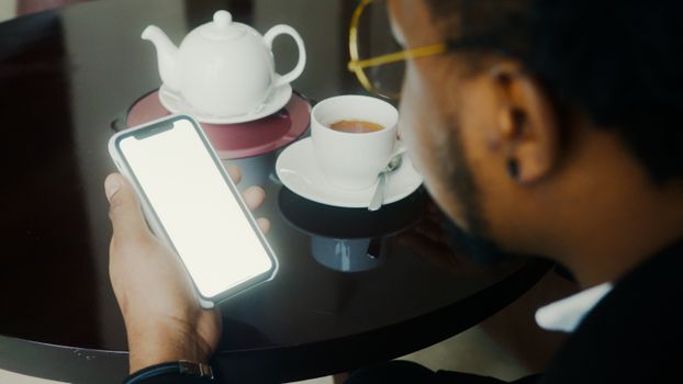Close-up of a man using a smartphone while enjoying tea indoors.