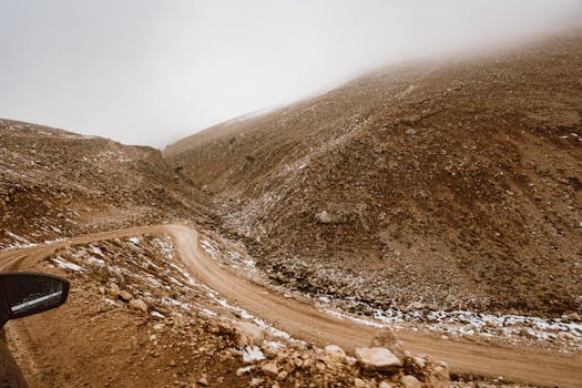A twisting dirt road winds through a foggy, mountainous terrain blanketed in light snow.